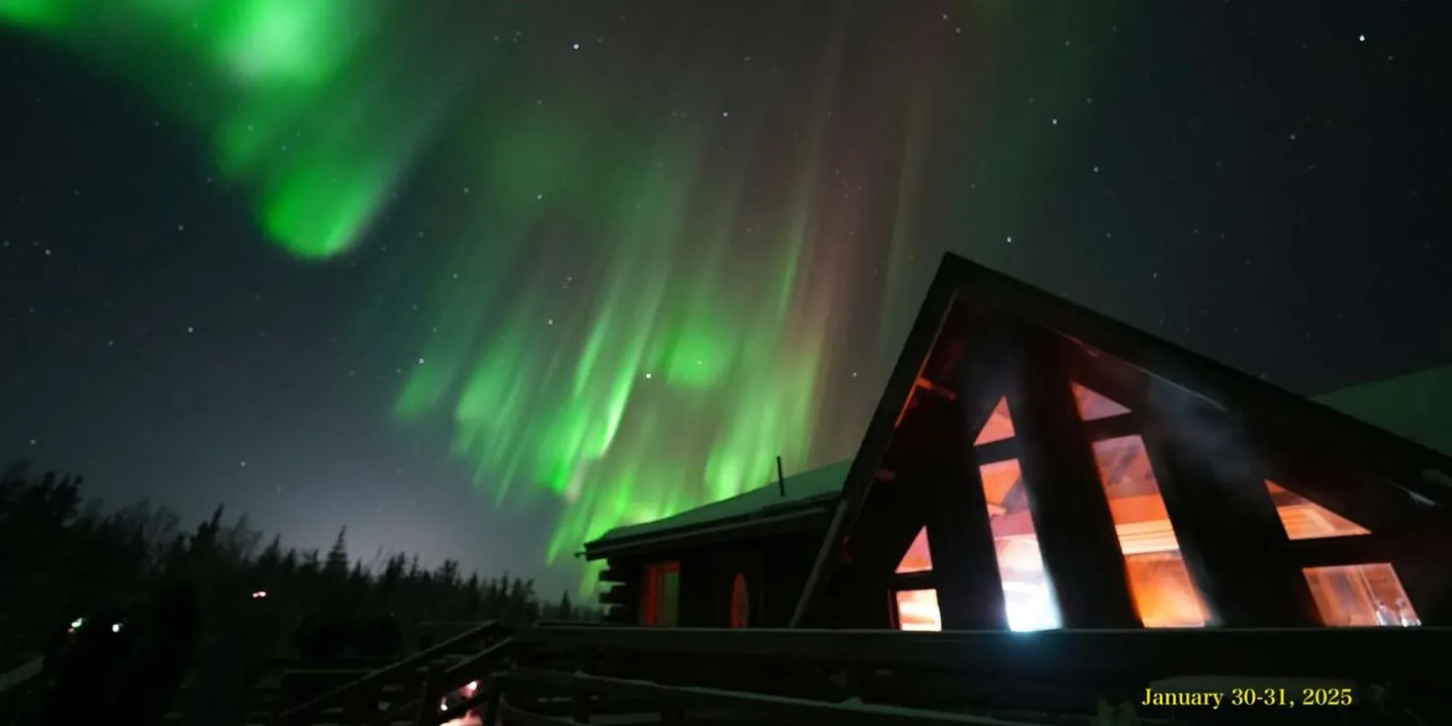 A-frame lodge cabin glowing orange from inside under a green aurora display