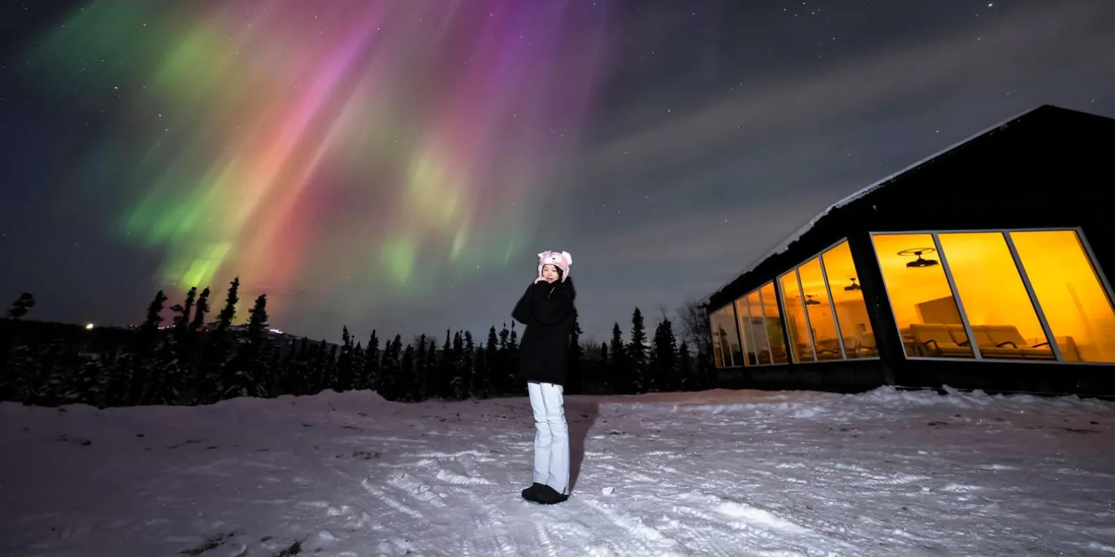 Guest under a pink and green aurora beside the warmly lit Aurora Glass House at Cleary Summit