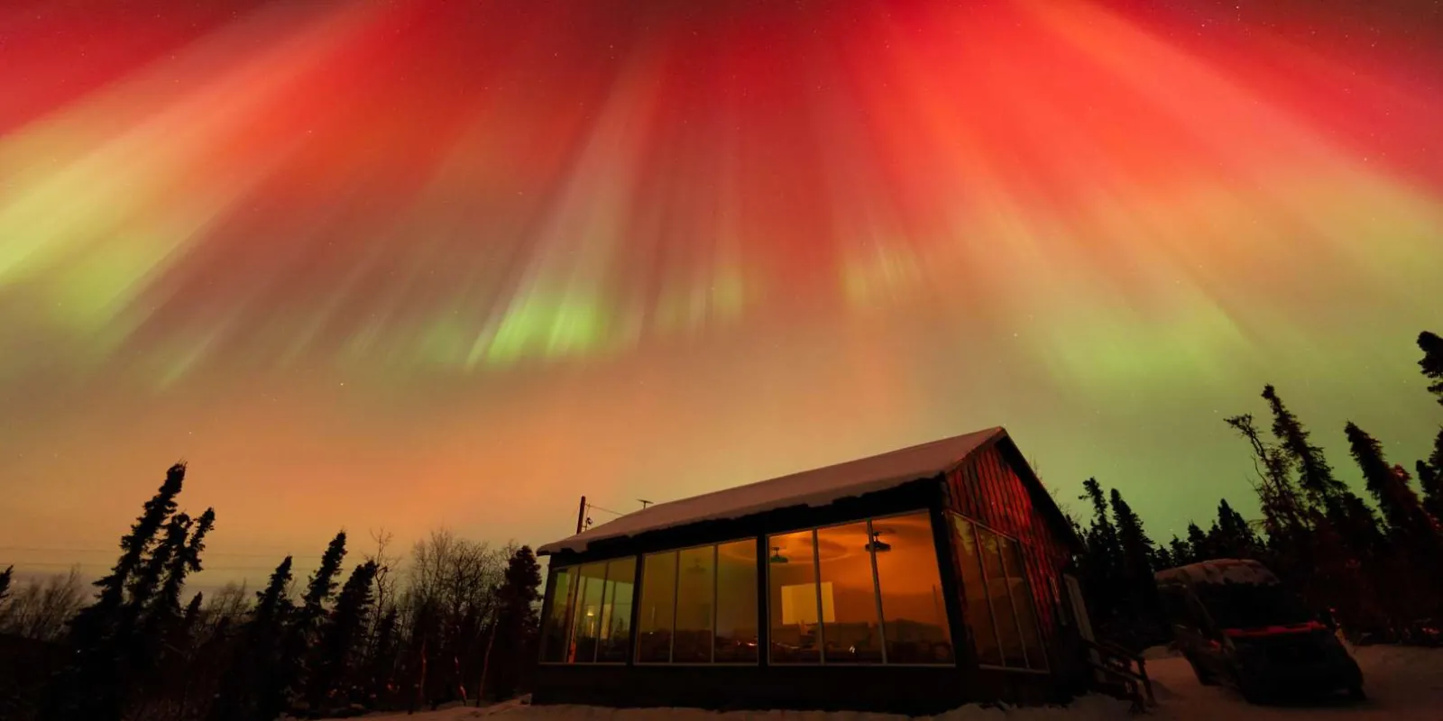 The glass-fronted Aurora Glass House cabin lit from inside under a red and green aurora