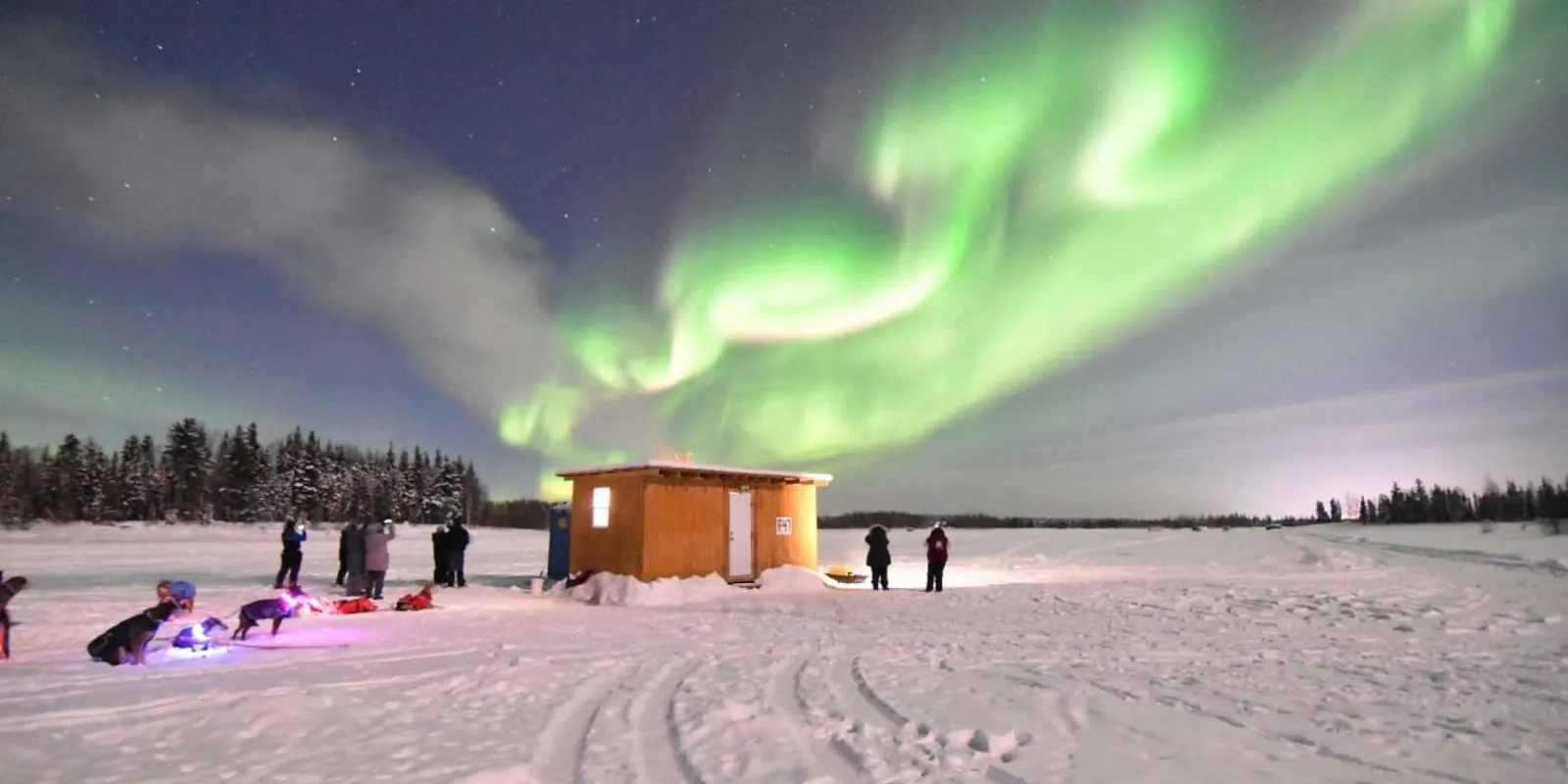 Green aurora over an ice fishing cabin and parked dog teams on a frozen lake near North Pole