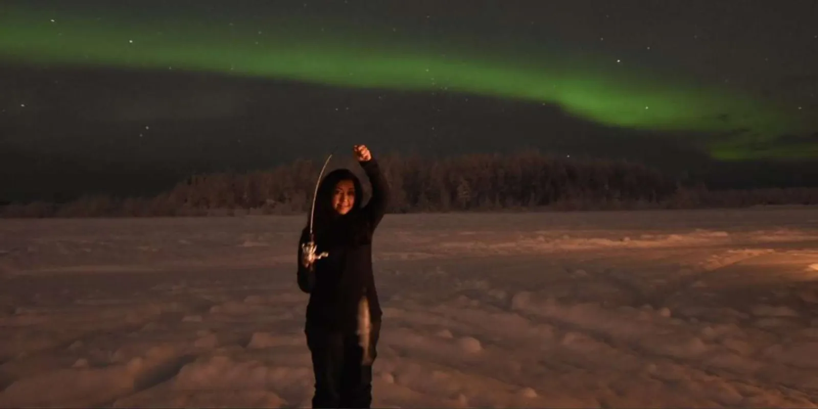 Guest on a frozen lake at night with a green aurora arching across the sky