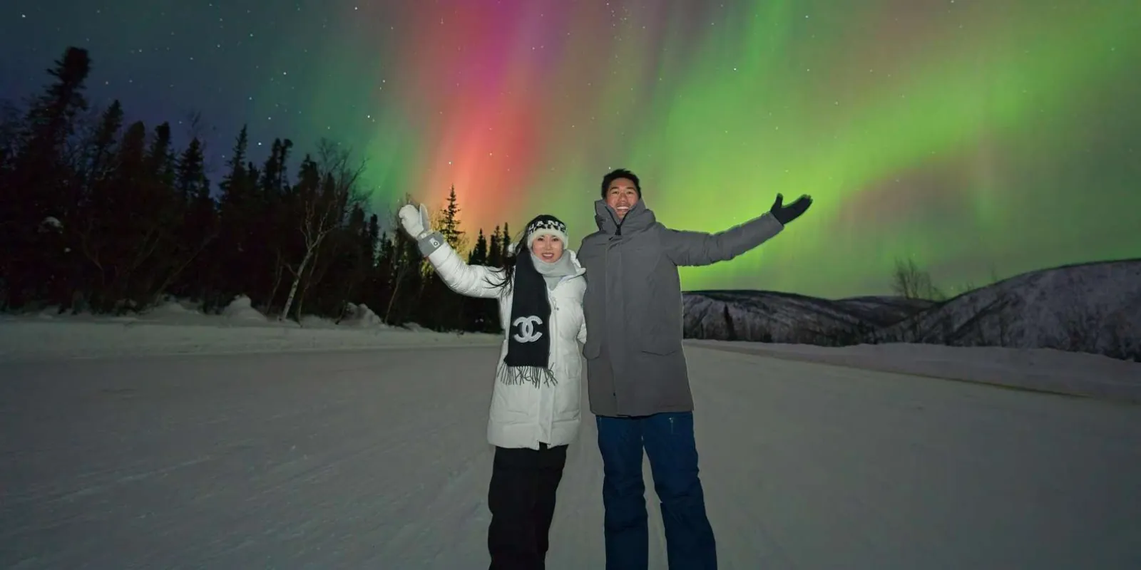 Couple with arms raised under green, violet, and pink aurora on a snow-covered Fairbanks road