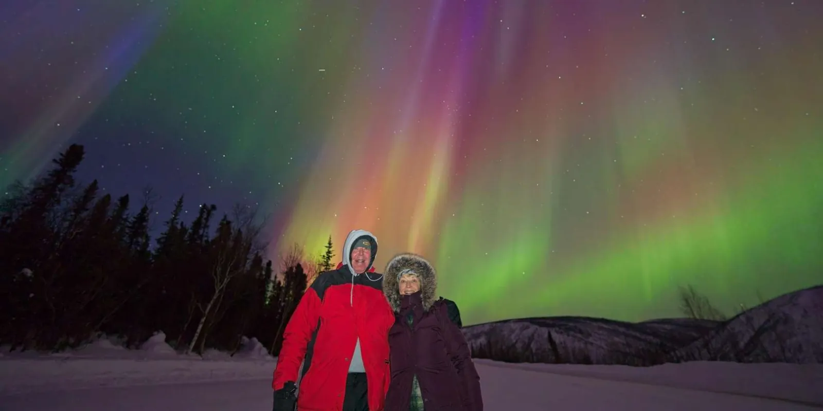 Guests in red and purple winter coats smiling under a pink and green aurora display