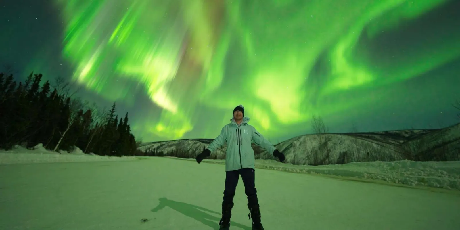 Solo traveler with arms outstretched beneath a vivid green aurora over a frozen valley