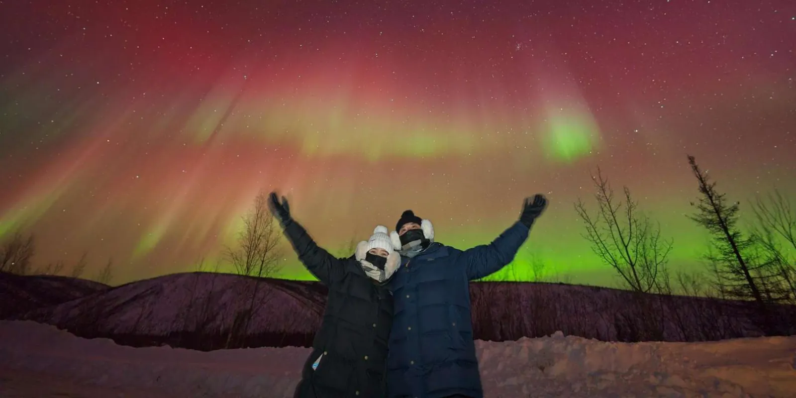 Couple with arms raised cheering under a deep red and green aurora curtain