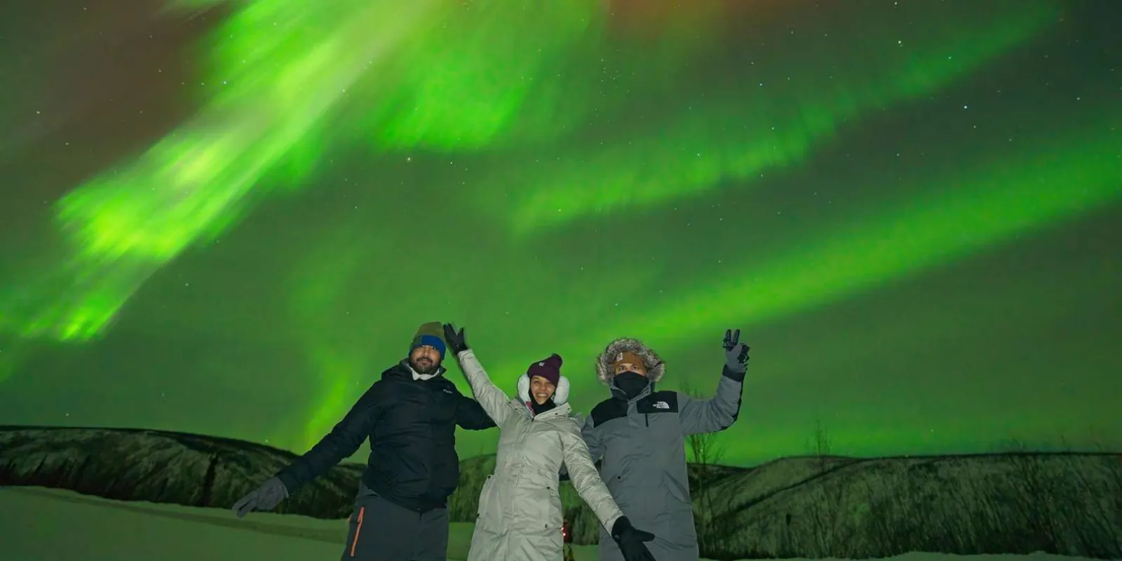 Group of three friends celebrating under a bright green aurora display near Fairbanks