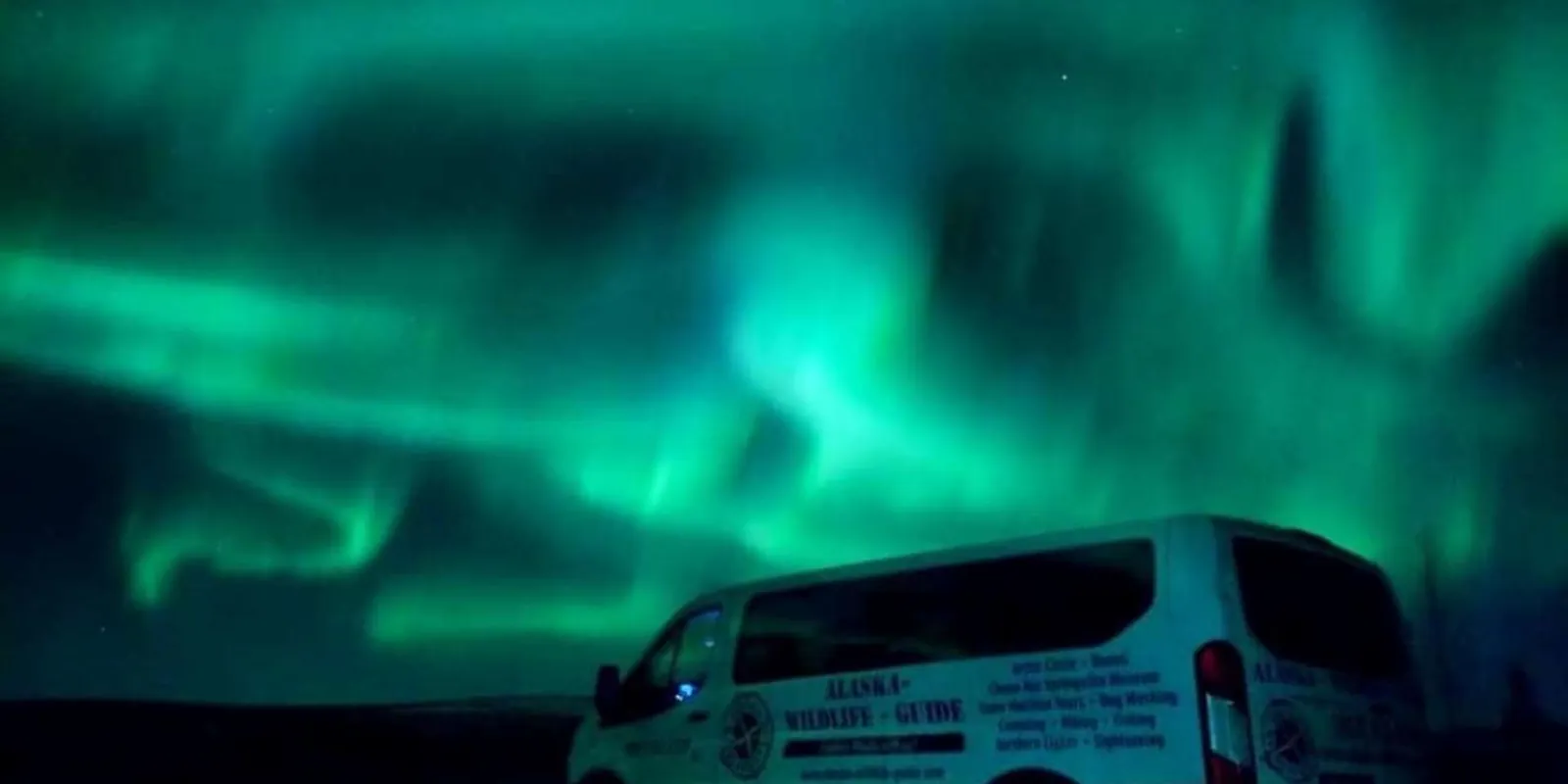Green aurora glowing above an Alaska Wildlife Guide tour van parked on the highway
