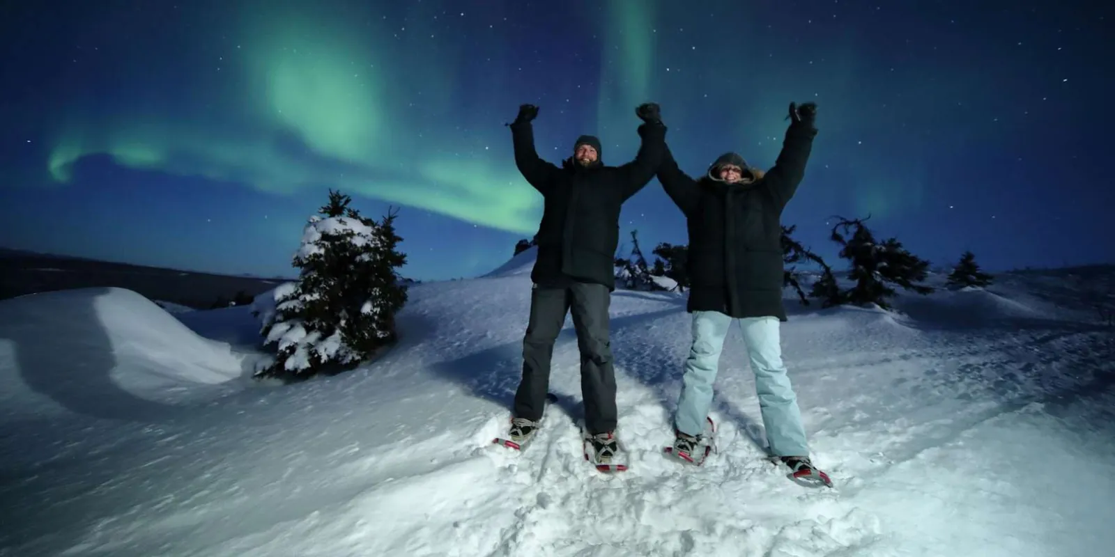Couple in snowshoes celebrating with raised arms under a green aurora display